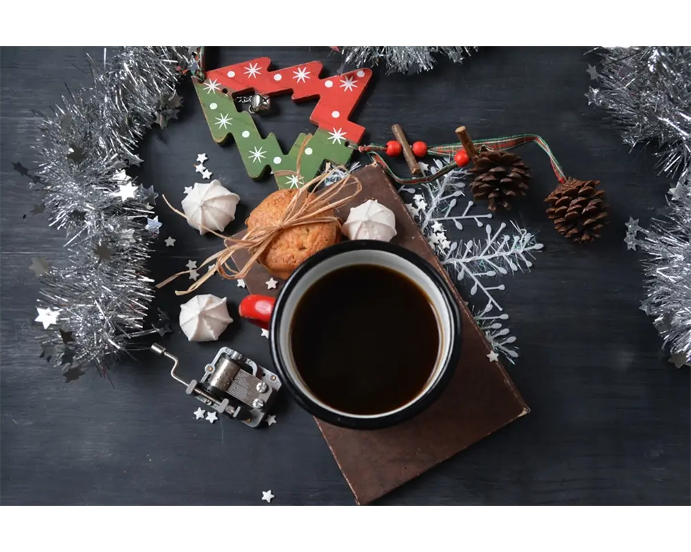 Et festlig høytidsarrangement med en kopp julekaffe, småkaker, en reinsdyrdekorasjon, kongler og sølvglitter på en mørk treflate.